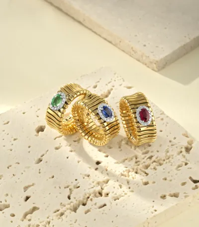 Three yellow gold Tubogas rings with oval green, blue and red gemstones in diamond halos