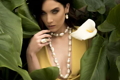 Model wearing a long baroque pearl necklace, pearl earrings and ring among green leaves